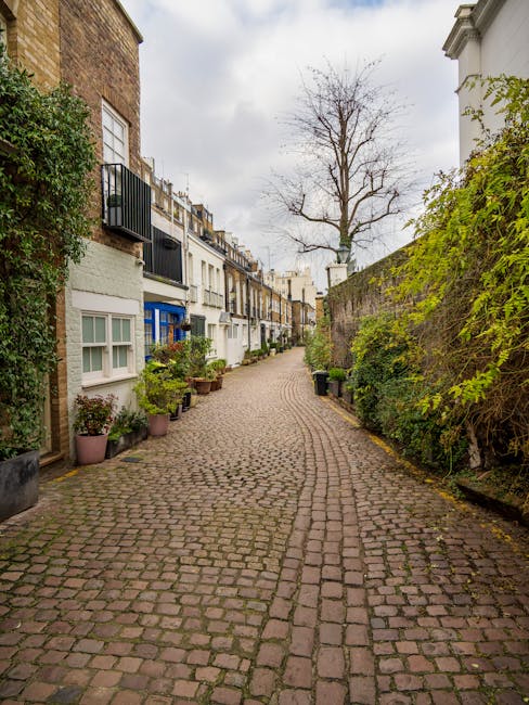 A narrow cobblestone street in South Kensington lined with terraced houses featuring small front gardens and potted plants. The houses have white or light-colored facades, some with balconies and black railings. A large, leafless tree stands in the background under an overcast sky. On the right side, a tall stone wall covered with green foliage runs along the sidewalk. The street appears quiet and residential, with no visible vehicles or pedestrians. This setting exemplifies a typical London neighbourhood suitable for house removals and relocation services, as provided by Man and Van South Kensington, who specialise in careful furniture transport and packing during home moves.