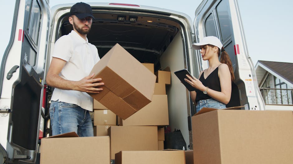 A man and a woman are working together during a home relocation, standing at the open rear of a white moving van parked outside a residential property. The man, dressed in a white polo shirt, jeans, and a black cap, is holding a medium-sized cardboard box, preparing to load or unload it. Inside the van, several other cardboard boxes of various sizes are stacked, some wrapped in plastic or tissue paper, indicating packing materials used for furniture transport or packing during house moves. The woman, wearing a black sleeveless top, a white cap, and jeans, is focused on a tablet device, possibly coordinating or checking inventory. The scene is set in natural daylight, with a pavement and neighboring houses visible in the background, illustrating the loading process typical in professional removals for South Kensington. Man and Van South Kensington facilitates efficient packing and moving services, including the careful handling of boxes and furniture during the loading process for home relocations.