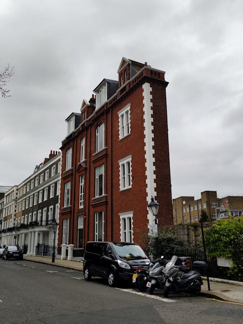 A multi-storey residential building made of red brick with white window frames and decorative vertical white accents along the corner, located on a city street under an overcast sky. In front of the building, a black van and three motorbikes are parked on the pavement, with some green shrubbery visible behind a black fence. The scene is typical of London architecture, and the image relates to home relocation or moving logistics, which is relevant to Man and Van South Kensington's removals services.