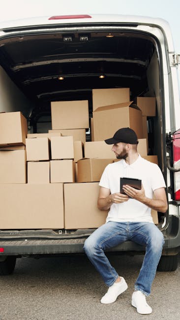 A man and a woman are working together during a home relocation, standing at the open rear of a white moving van parked outside a residential property. The man, dressed in a white polo shirt, jeans, and a black cap, is holding a medium-sized cardboard box, preparing to load or unload it. Inside the van, several other cardboard boxes of various sizes are stacked, some wrapped in plastic or tissue paper, indicating packing materials used for furniture transport or packing during house moves. The woman, wearing a black sleeveless top, a white cap, and jeans, is focused on a tablet device, possibly coordinating or checking inventory. The scene is set in natural daylight, with a pavement and neighboring houses visible in the background, illustrating the loading process typical in professional removals for South Kensington. Man and Van South Kensington facilitates efficient packing and moving services, including the careful handling of boxes and furniture during the loading process for home relocations.
