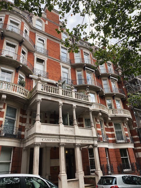 A multi-storey red-brick building with ornate white stone detailing and balconies on each floor, situated on Campden Hill Court. The building features curved bay windows and a prominent entrance supported by classical columns, with a rounded porch area. Overhanging tree branches with green leaves partially obscure the upper part of the building, and parked cars are visible along the street at the base. The image captures a typical London residential facade, relevant to home relocation and furniture transport services, as seen in the context of Cromwell Road same-day removals in South Kensington operated by Man and Van South Kensington.