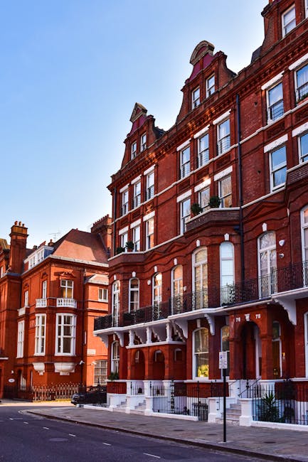 The image shows a multi-storey red brick building with large sash windows and decorative iron balconies lining the front facade, situated on Gloucester Road in South Kensington. The building features a prominent arched entrance with white trim and steps leading up to the doorway. In the foreground, a pavement runs along the street, which is clear of traffic and lined with pigeon droppings. A white van with a sliding side door is parked on the street, with its rear door open during a house removal or relocation process. Inside the van, seen through the open door, are several cardboard boxes, some wrapped in plastic or protective packaging, and a few rolled-up carpets or blankets used for furniture protection. Nearby, a person wearing gloves is in the process of lifting or carrying a box towards the van, indicating active loading and packing for a home relocation. The scene is set during daylight, with bright blue sky overhead, and the overall environment reflects a professional moving service by Man and Van South Kensington, specializing in removals and furniture transport in the area.