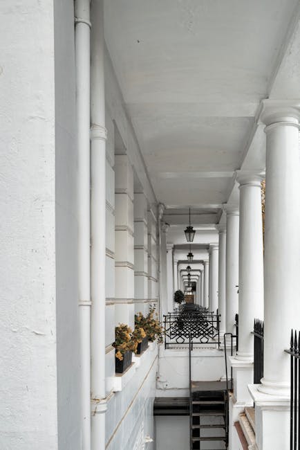 The image shows a multi-storey red brick building with large sash windows and decorative iron balconies lining the front facade, situated on Gloucester Road in South Kensington. The building features a prominent arched entrance with white trim and steps leading up to the doorway. In the foreground, a pavement runs along the street, which is clear of traffic and lined with pigeon droppings. A white van with a sliding side door is parked on the street, with its rear door open during a house removal or relocation process. Inside the van, seen through the open door, are several cardboard boxes, some wrapped in plastic or protective packaging, and a few rolled-up carpets or blankets used for furniture protection. Nearby, a person wearing gloves is in the process of lifting or carrying a box towards the van, indicating active loading and packing for a home relocation. The scene is set during daylight, with bright blue sky overhead, and the overall environment reflects a professional moving service by Man and Van South Kensington, specializing in removals and furniture transport in the area.