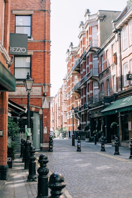 The image depicts a narrow cobblestone street in South Kensington, with multistory brick buildings lining both sides. On the left, a brick building with a dark green sign reading 'HEZZO' is visible near a vintage-style lamppost. The street appears to be part of a commercial area, with storefronts and awnings extending from the ground floors of the buildings. In the foreground, black bollards are positioned along the pavement, guiding pedestrian movement. The scene is well-lit with natural daylight, and the perspective leads toward a gently curving street that narrows in the distance. The overall atmosphere is quiet and typical of an urban residential and shopping district, suitable for home relocation activities or furniture transport planning by companies like Man and Van South Kensington, as the environment reflects an area where moving and packing services are often required.