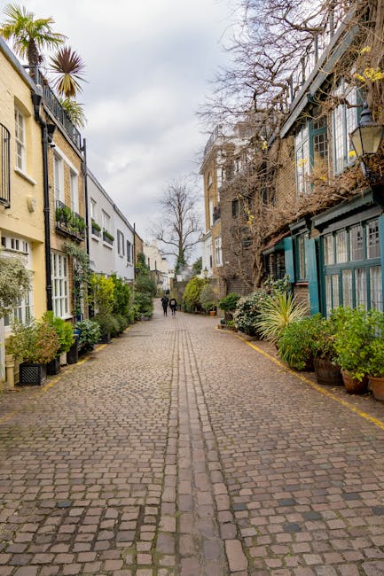 A cobblestone street lined with multi-storey residential buildings on both sides, featuring a mix of modern and traditional architectural styles. The buildings are painted in light colors, with some having balconies and large windows. Potted plants and greenery are visible along the pavement, with taller trees and shrubs interspersed among the buildings. The street appears quiet, with a few pedestrians walking in the distance. The sky is cloudy, providing diffused lighting. In the context of house removals, this setting reflects a typical urban environment where furniture and packing materials may be transported for home relocation services by companies such as Man and Van South Kensington, with logistics managed within narrow street environments.
