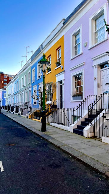 A row of colorful terraced houses along a paved South Kensington street, with pastel facades in shades of blue, yellow, purple, and white. Each house has a small front garden with black wrought iron railings and steps leading up to white doorways. A traditional black lamppost is positioned near the pavement, and some houses feature small window boxes with plants. The sky above is clear and blue, indicating a bright, sunny day. The street appears quiet with no visible pedestrians or vehicles. The scene captures the typical residential architecture of South Kensington, reflecting a well-maintained, desirable neighbourhood. This image is relevant to [COMPANY_NAME]’s house removals services focused on home relocation, furniture transport, and packing and moving activities in the South Kensington area.