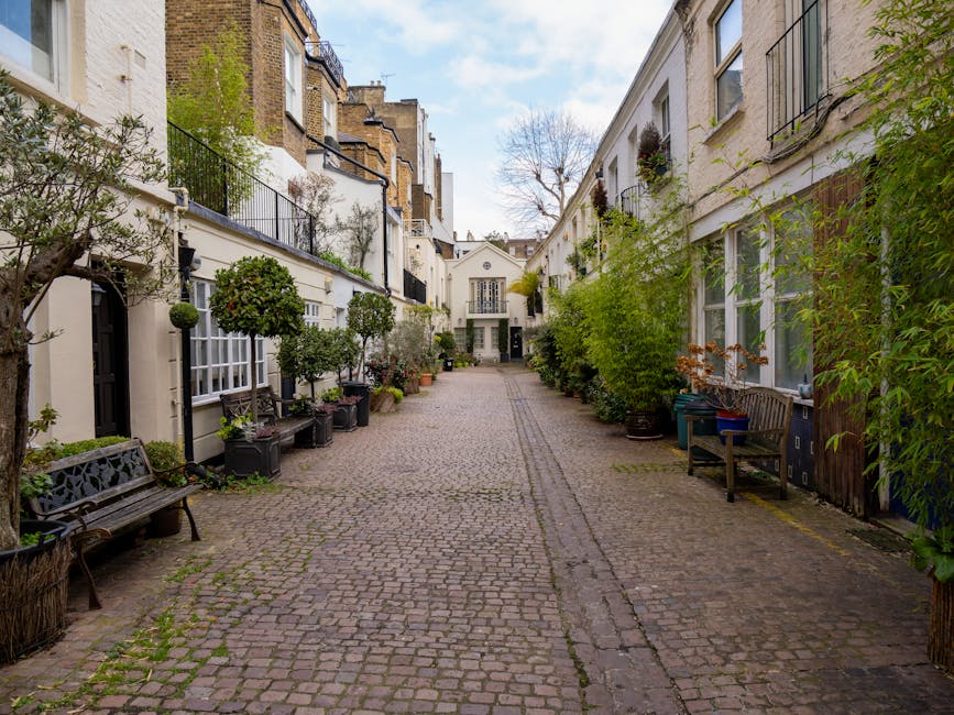 A cobblestone street lined with multi-storey residential buildings on both sides, featuring a mix of modern and traditional architectural styles. The buildings are painted in light colors, with some having balconies and large windows. Potted plants and greenery are visible along the pavement, with taller trees and shrubs interspersed among the buildings. The street appears quiet, with a few pedestrians walking in the distance. The sky is cloudy, providing diffused lighting. In the context of house removals, this setting reflects a typical urban environment where furniture and packing materials may be transported for home relocation services by companies such as Man and Van South Kensington, with logistics managed within narrow street environments.